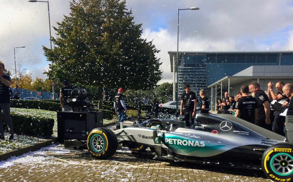 Mercedes F1 team celebrates their world championship victory at the factory with Toto Wolff and Niki Lauda present, Silver Arrows car front and center, white confetti cannons creating a celebratory atmosphere with crew members in the background.