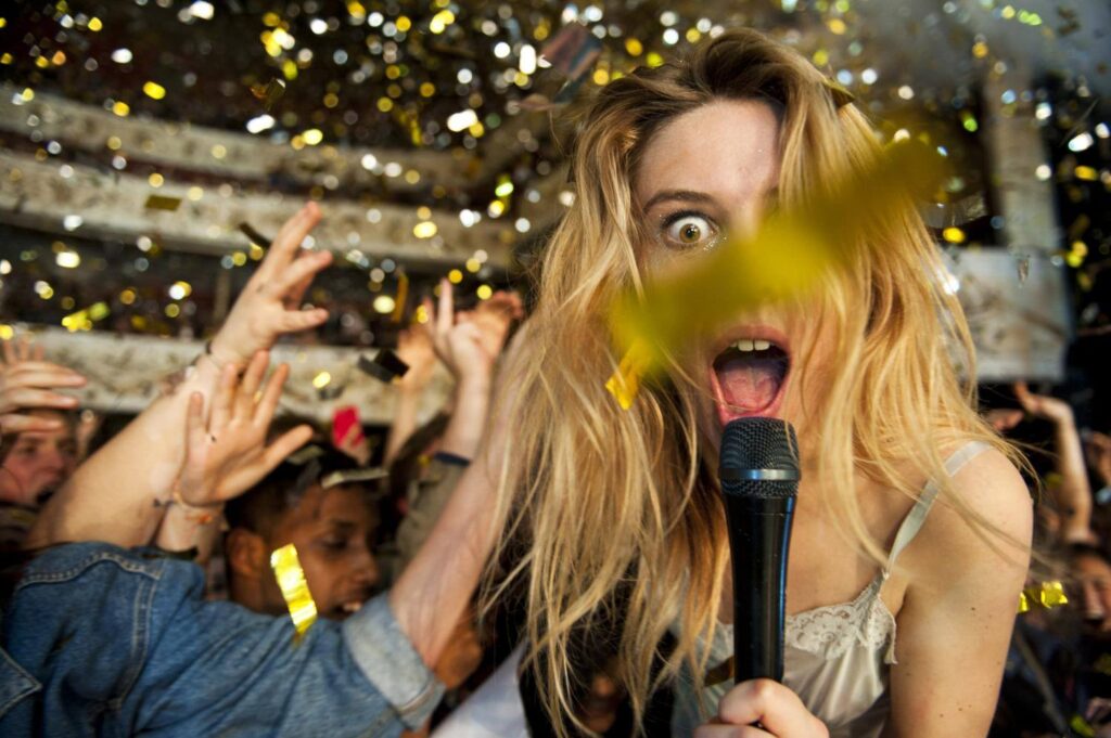 Ellie Rowsell from Wolf Alice performing at Shepherd's Bush Empire with gold confetti cannons firing across the stage, three tiers of seating visible in the background, and confetti flowing over the crowd during the live concert.