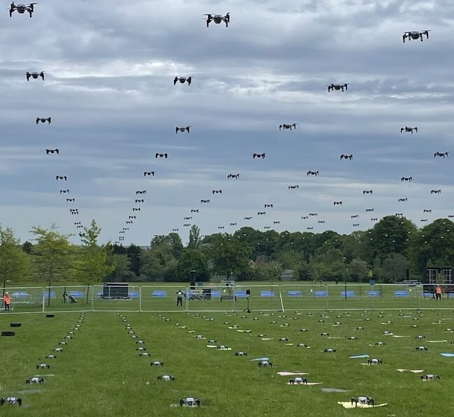 UK drone display: A daytime event with 100 drones in a precise grid formation flying above a field, and another 100 drones on the ground. The audience barrier and steward are visible in the background. This striking UK drone show highlights coordinated aerial displays in a public event.