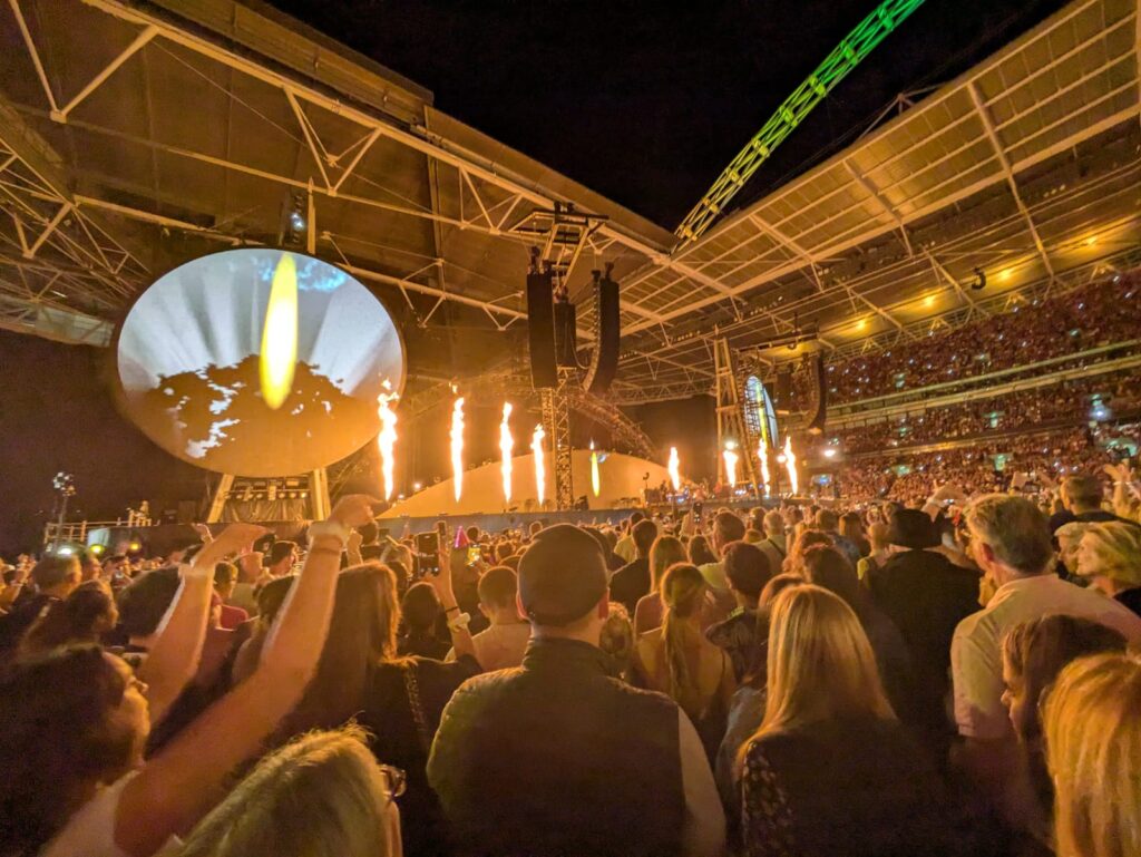 Coldplay performing at Wembley Stadium, with a sea of fans, the iconic Wembley arch, and flames on stage.