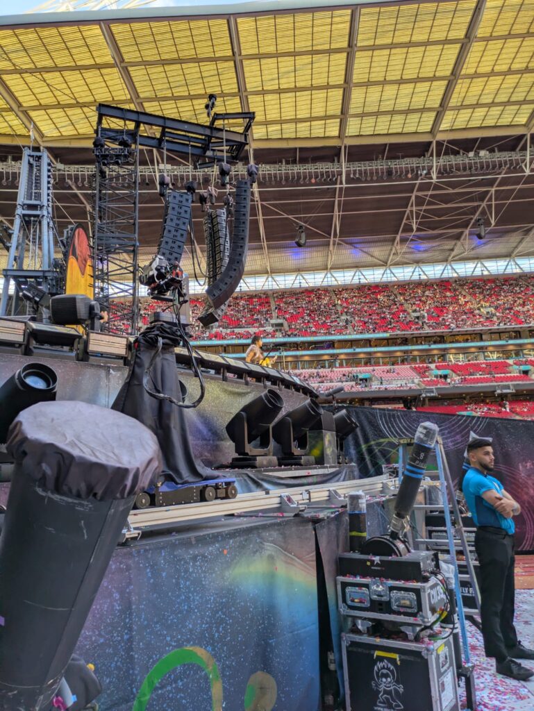 Wembley Stadium Coldplay concert infrastructure during the day, showing the stage setup, lighting rig, and seating from the side, along with a steward.