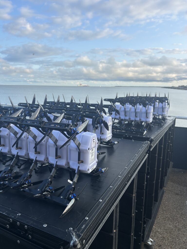 Fifteen drones stacked on their side atop black flight cases, with a bright blue sky in the background, as they prepare to be set up for a drone display.