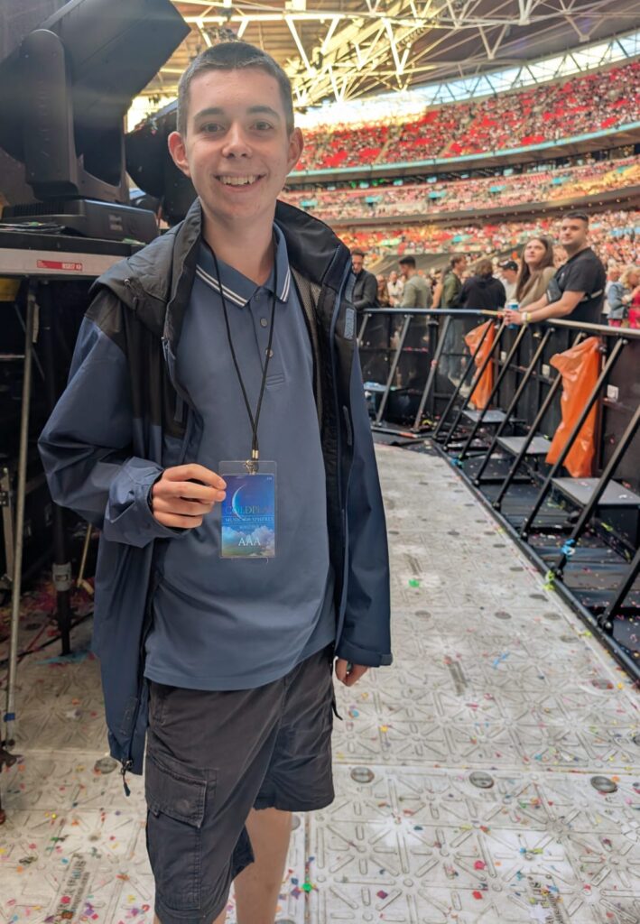 Wembley Stadium Coldplay concert: a young fan smiling with a backstage pass, standing by the stage barrier, with the stadium and walkway visible behind him during the day.