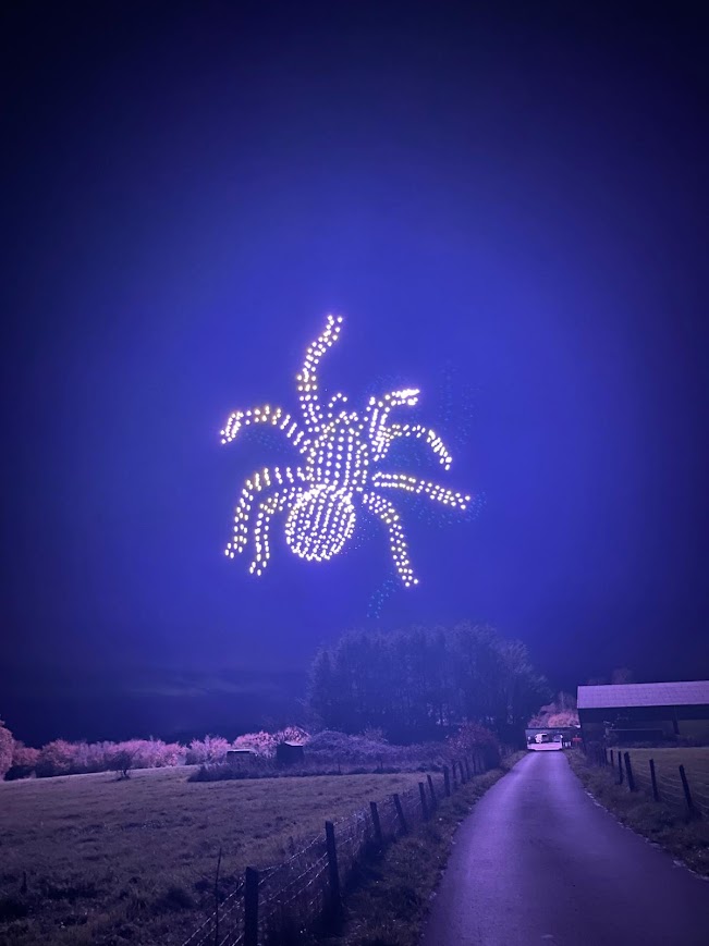 A drone formation shaped like a tarantula, glowing with a soft blue aura, hovering over a rural countryside field at night, surrounded by a deep blue sky.